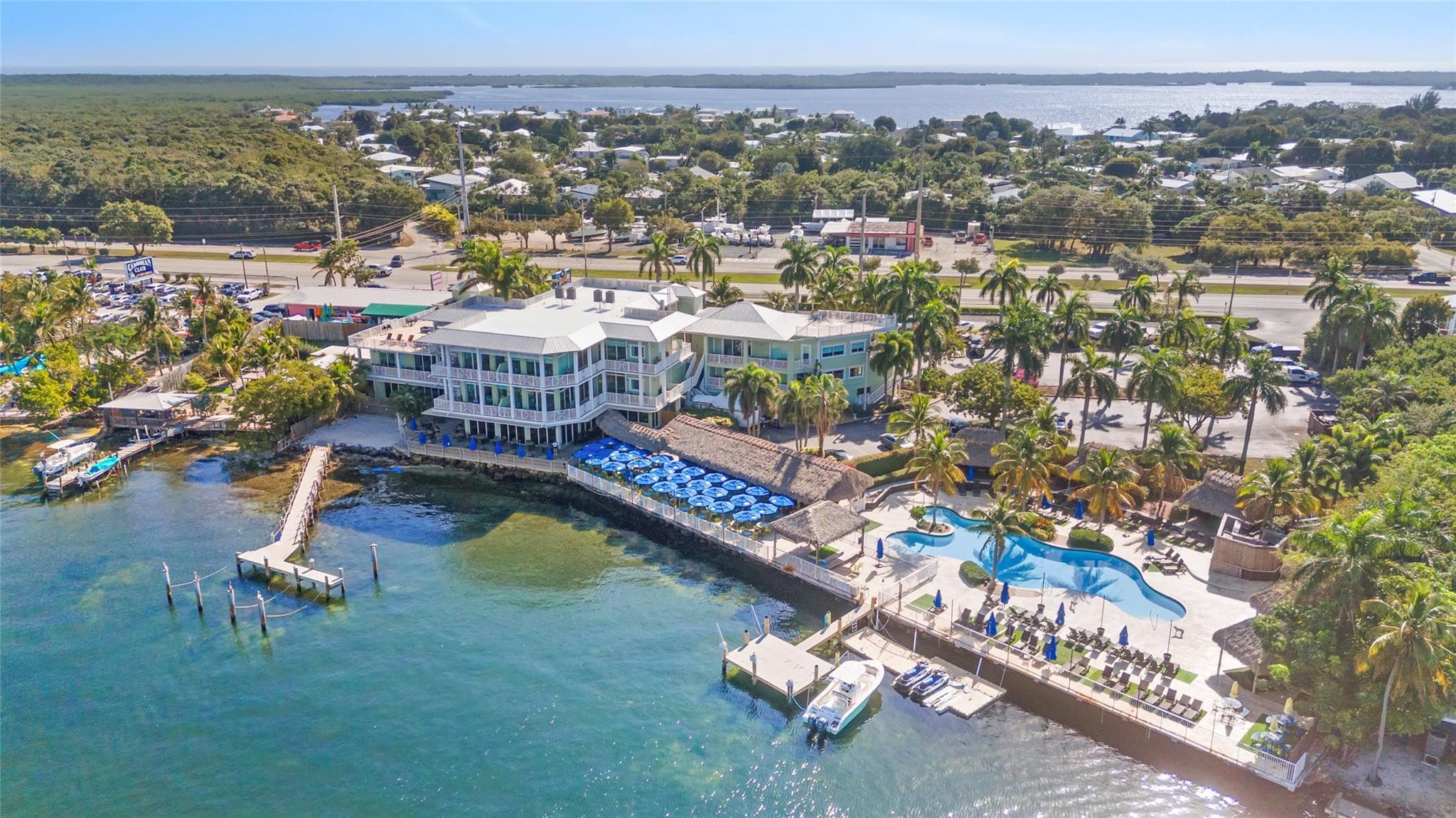 Aerial view of the bayfront residence club showing the white three-story building, free-form pool, thatched tiki huts, blue umbrellas, docks, and palm-lined grounds along the turquoise bay.