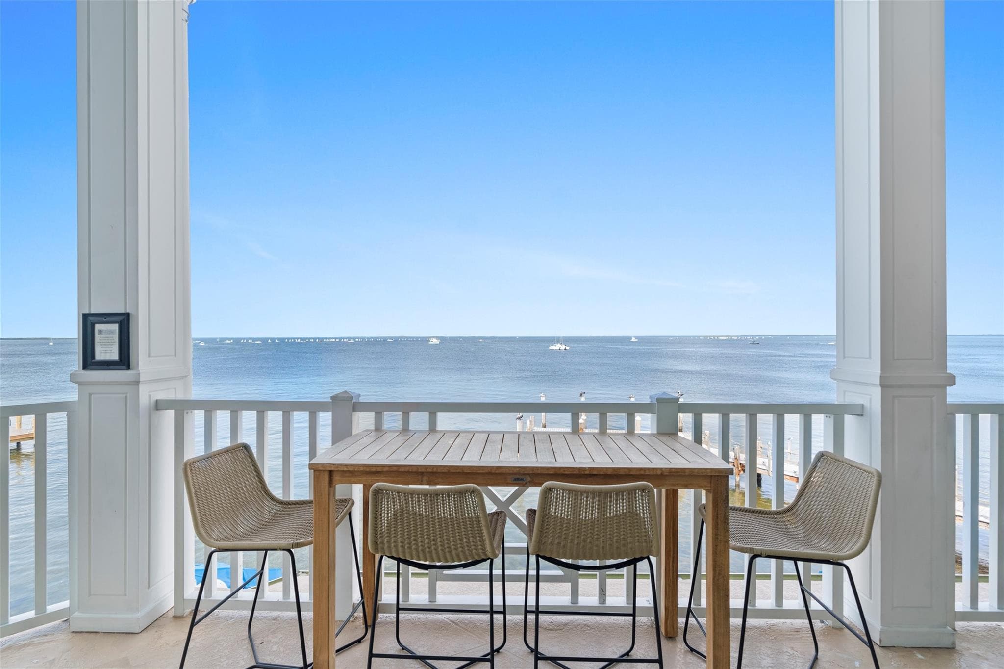 Teak bar-height table with four woven counter chairs on a columned balcony facing an unbroken horizon of calm bay water dotted with distant sailboats.