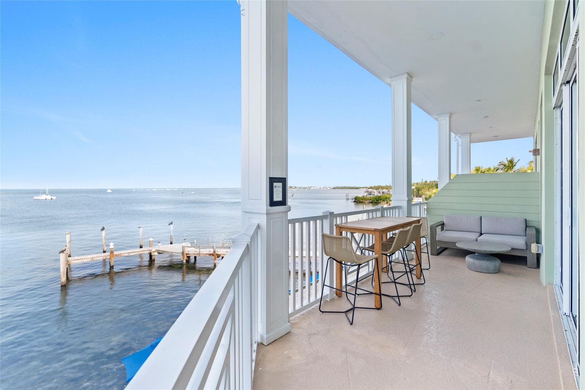 Covered balcony with white columns and railings, a teak bar-height table, wicker stools, and a gray outdoor sofa overlooking weathered dock pilings and calm bay water.