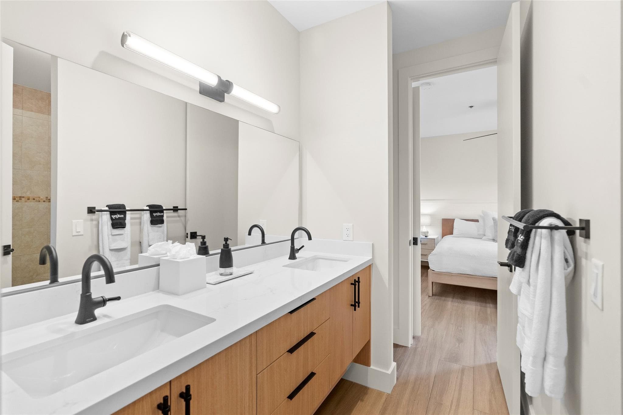 Long bathroom vanity with two undermount sinks, black fixtures, tube sconce lighting, and black-and-white towels, with a doorway opening to a bright bedroom.