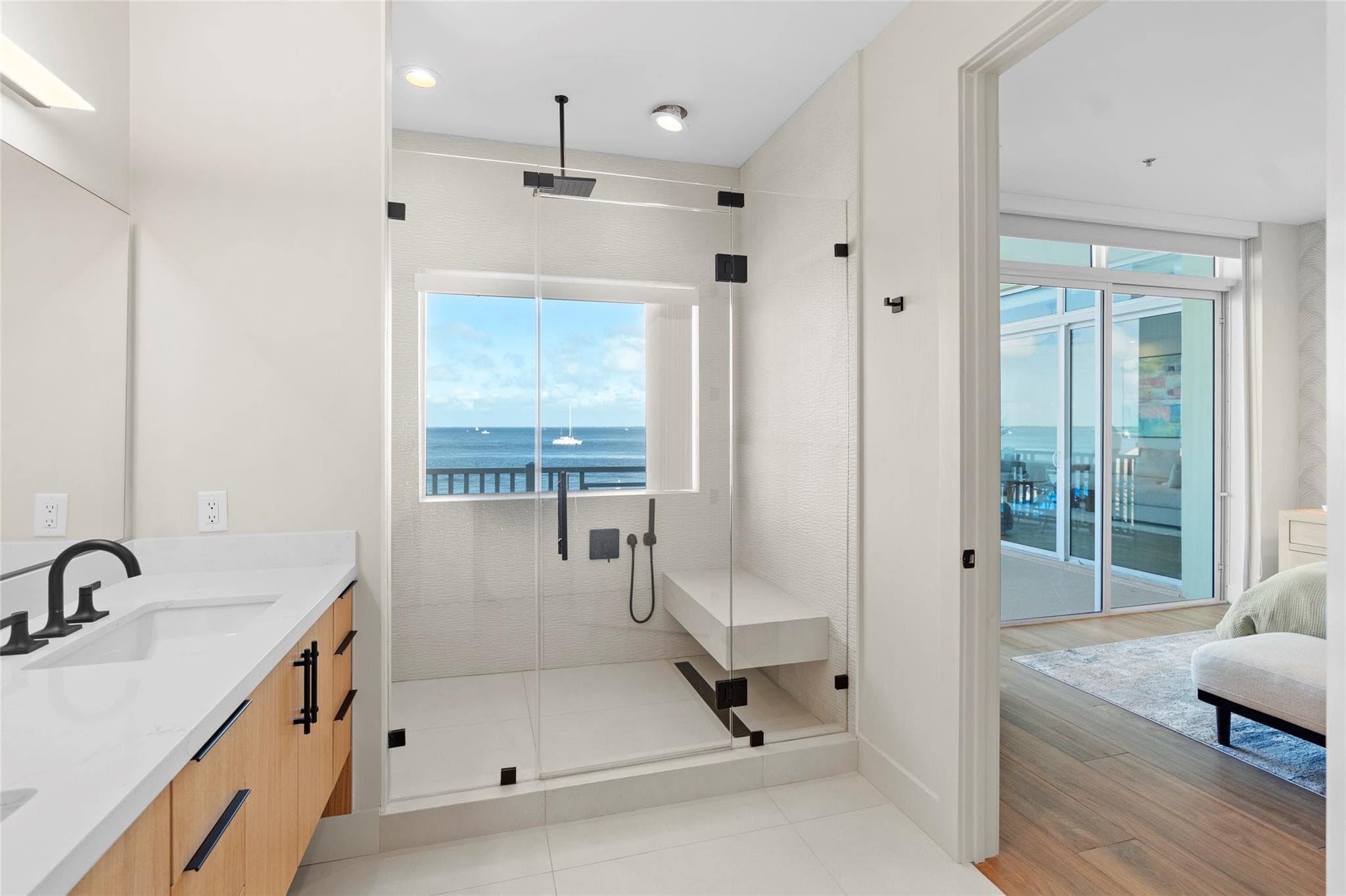 Glass-enclosed walk-in shower with rainfall head, floating bench, and a window framing the bay, beside a white quartz vanity with black faucet.