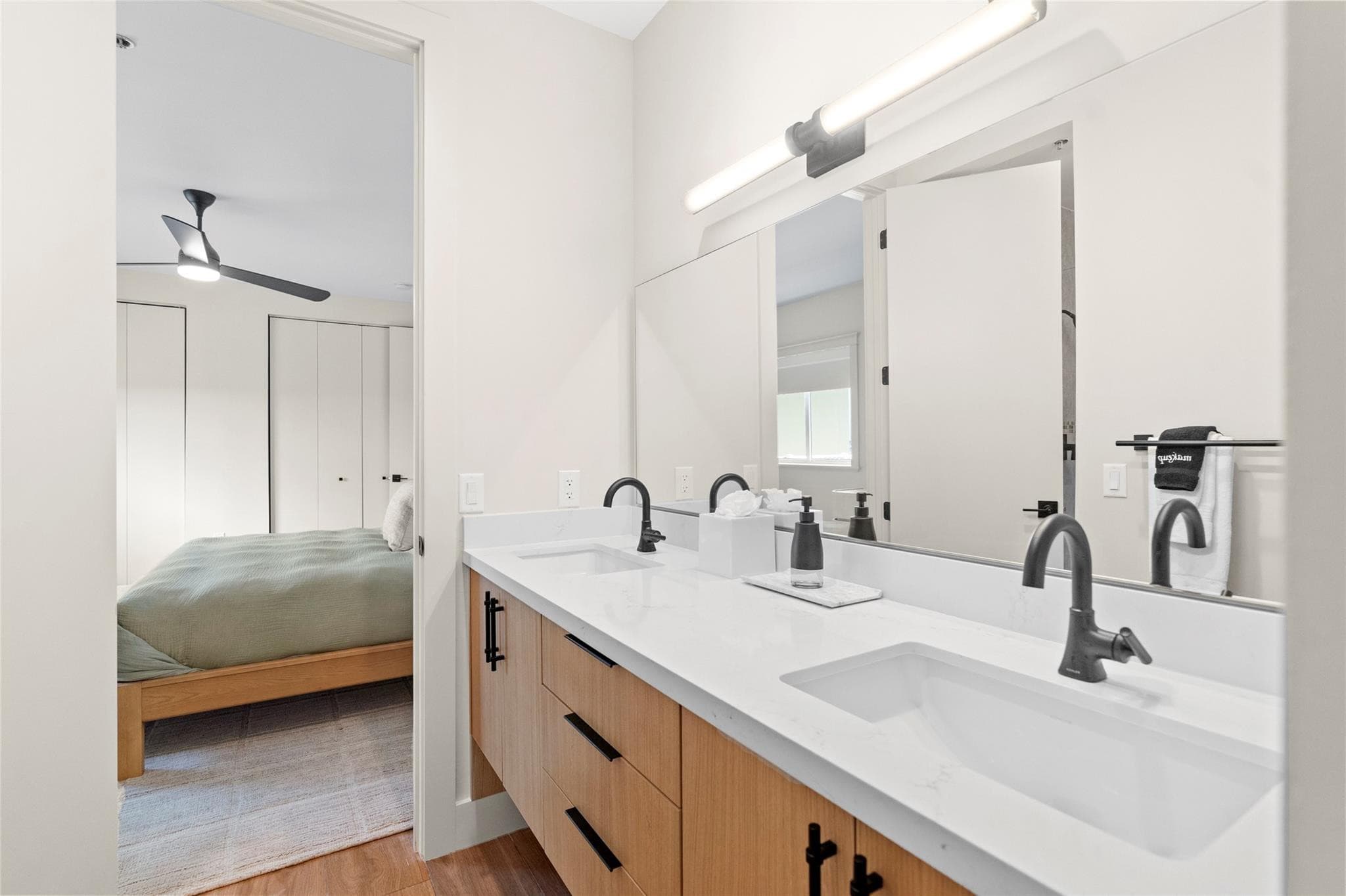 Bathroom with a wood double vanity, white quartz top, black gooseneck faucets, and an open door revealing the sage-green bed in the adjoining bedroom.