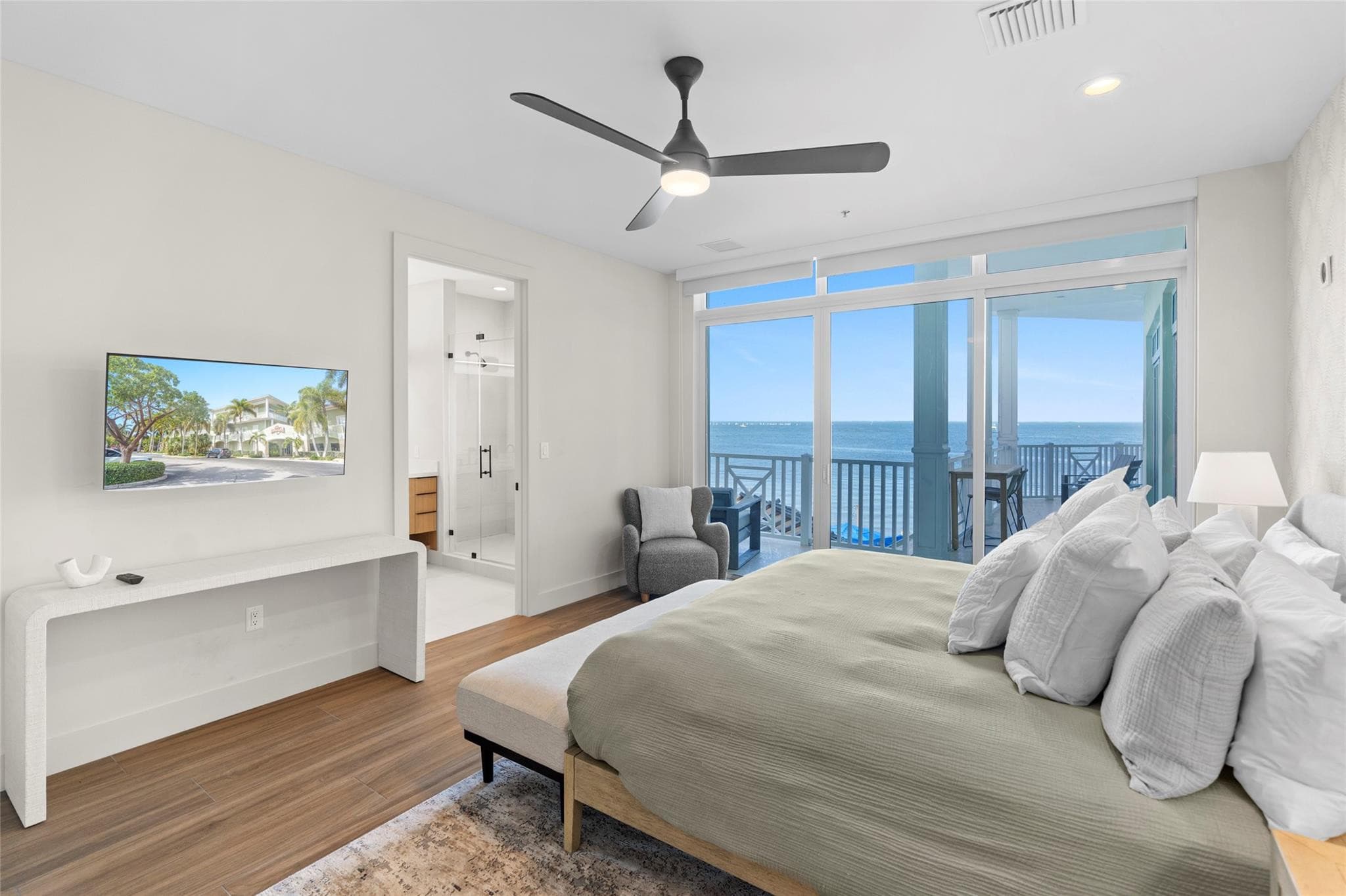 Bedroom with a wall-mounted TV, gray wing chair, en-suite bath, and glass sliders opening to a covered balcony with wide ocean views.