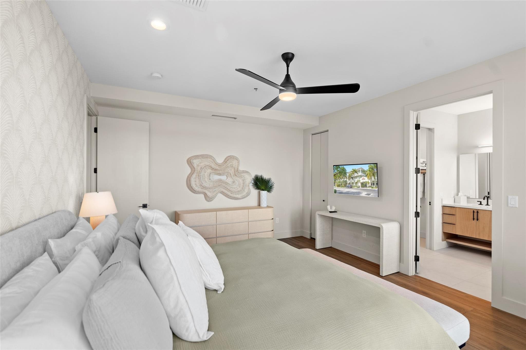 Neutral-toned bedroom with layered white pillows, a light-wood dresser under sculptural wall art, wall-mounted TV, and a doorway to the en-suite vanity.