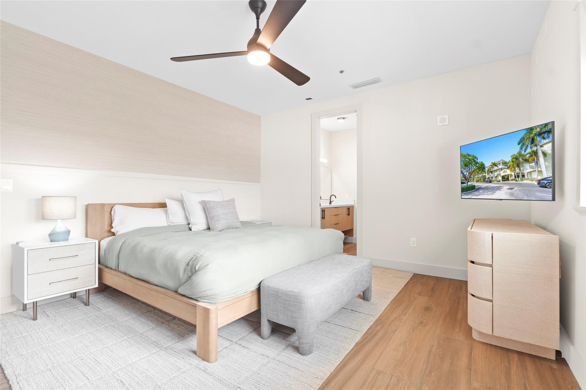 Serene bedroom with a light-wood bed, sage duvet, gray upholstered bench, woven dresser, ceiling fan, and an en-suite bathroom glimpsed through the doorway.
