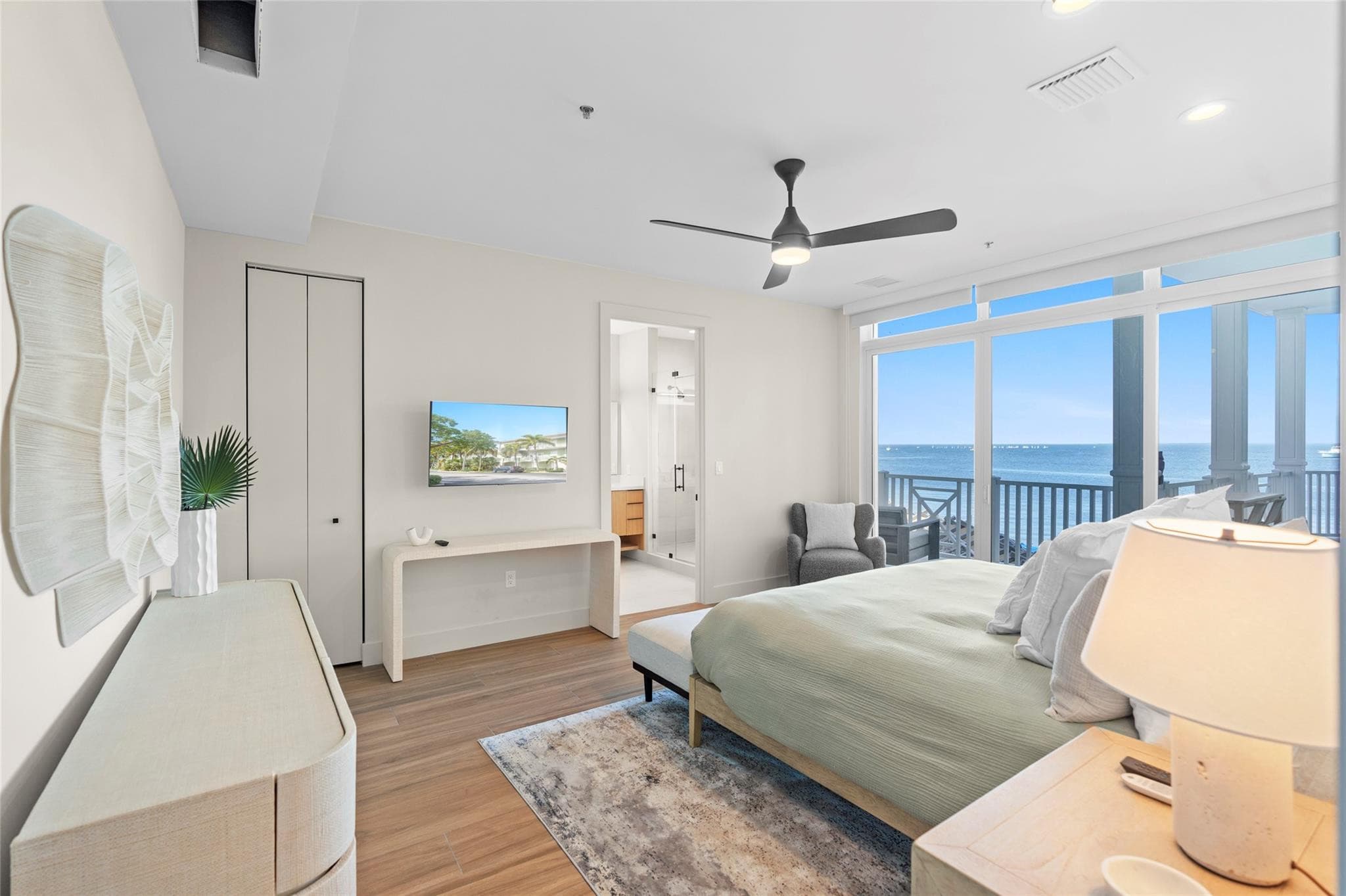 Spacious bedroom with woven wall art, a white console table with TV, en-suite bath, and floor-to-ceiling glass opening to a balcony over the open water.