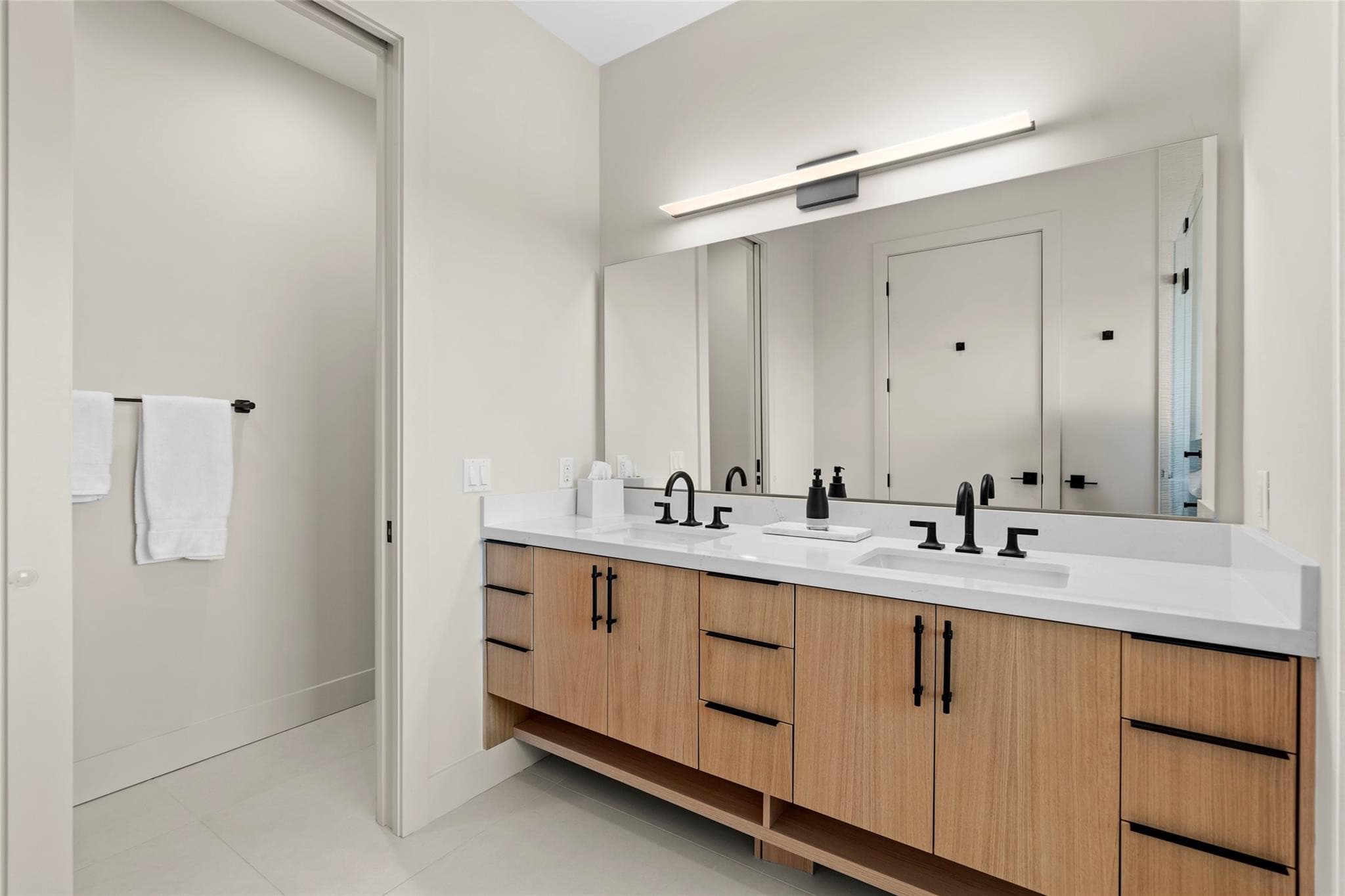 Bathroom with a long white-oak floating double vanity, white quartz counters, matte-black widespread faucets, and a full-width mirror under a linear sconce.