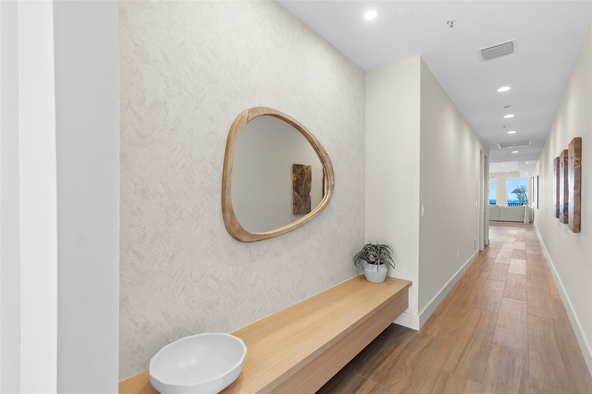 Entry hallway with a textured accent wall, organic-shaped wood-framed mirror, floating white-oak bench with a ceramic bowl, and wood-look tile leading to the water-view living area.