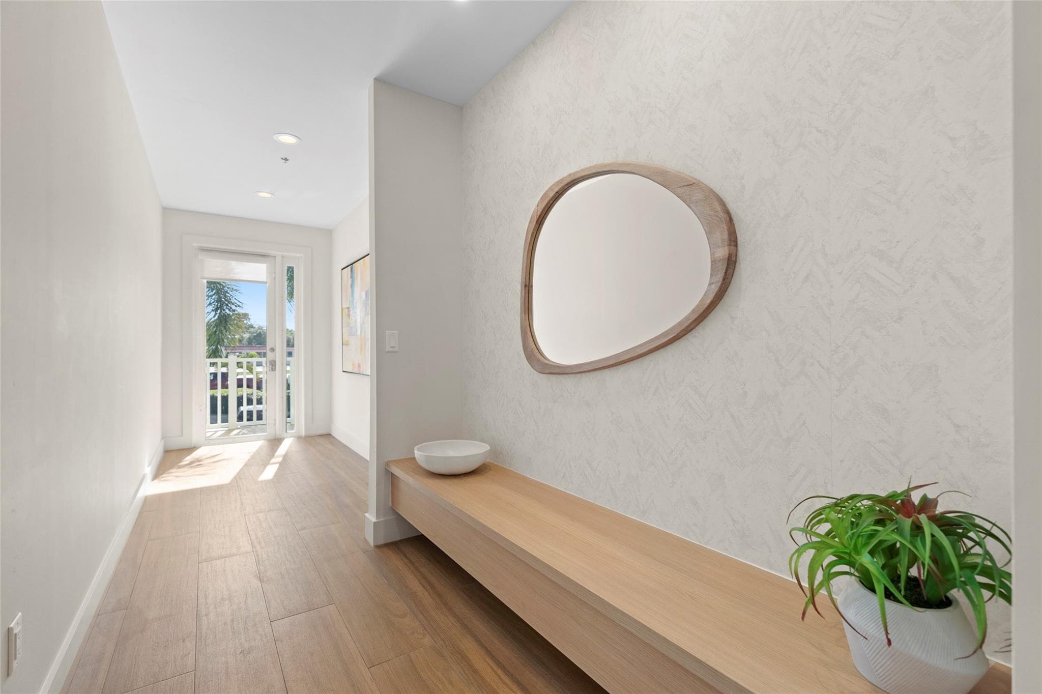 Bright foyer with a long floating wood bench, sculptural mirror on textured wallpaper, potted plant, and a glass French door letting in palm-filtered sunlight.