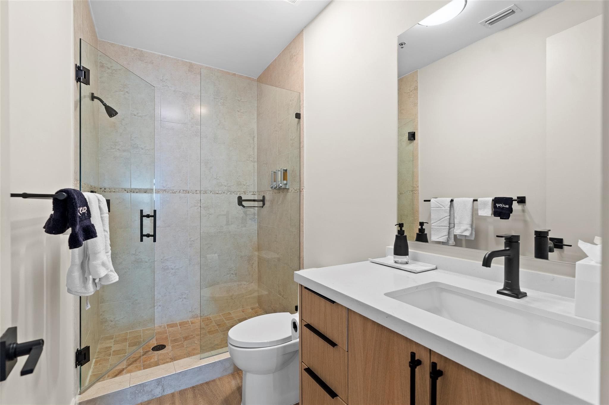 Guest bathroom with a frameless glass shower in warm beige tile with mosaic accent band, wood vanity with black faucet, and white quartz countertop.