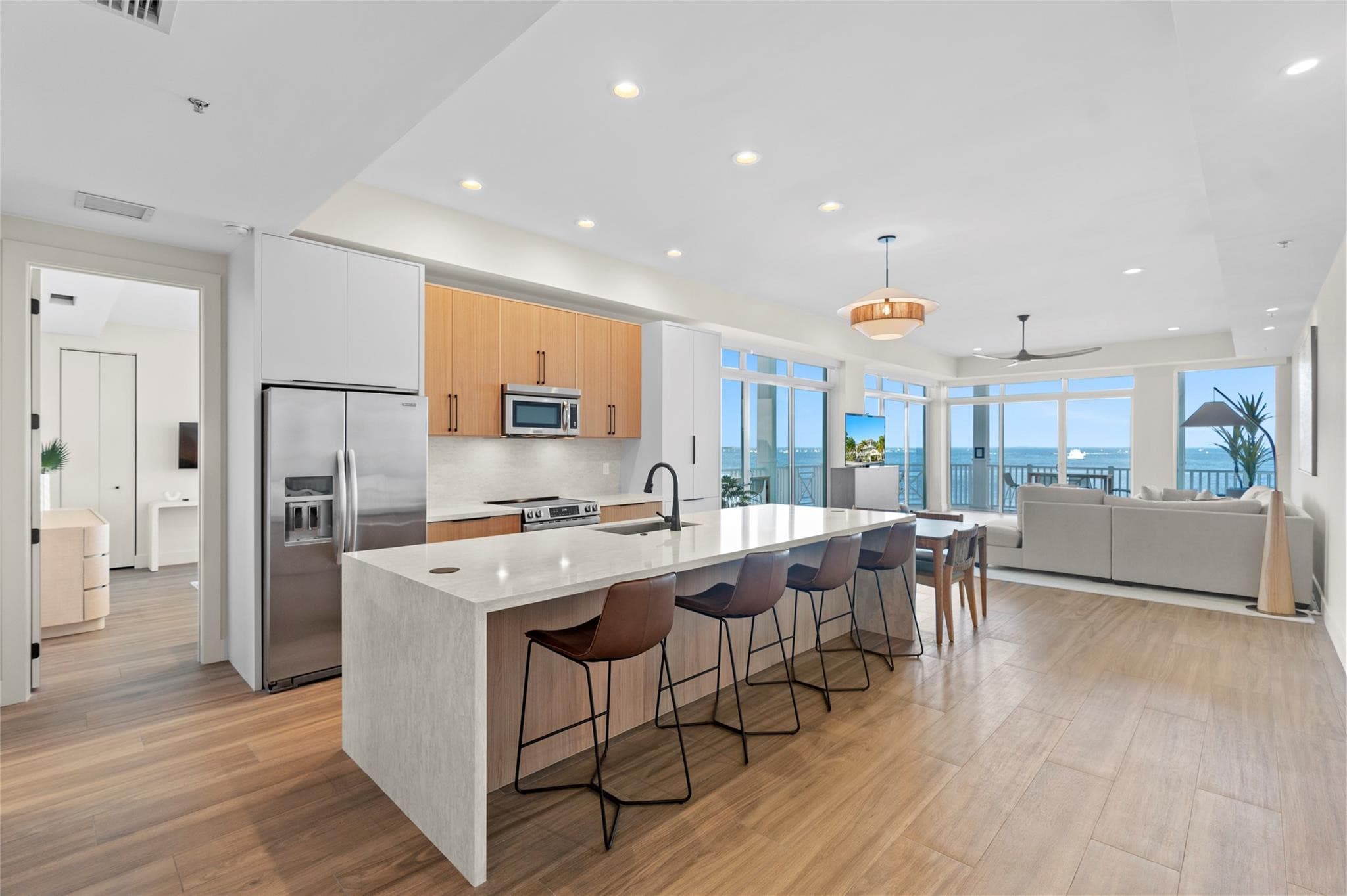 Open-plan kitchen with a long quartz waterfall island, four leather barstools, stainless appliances, and wood cabinetry opening to a living area with floor-to-ceiling water views.