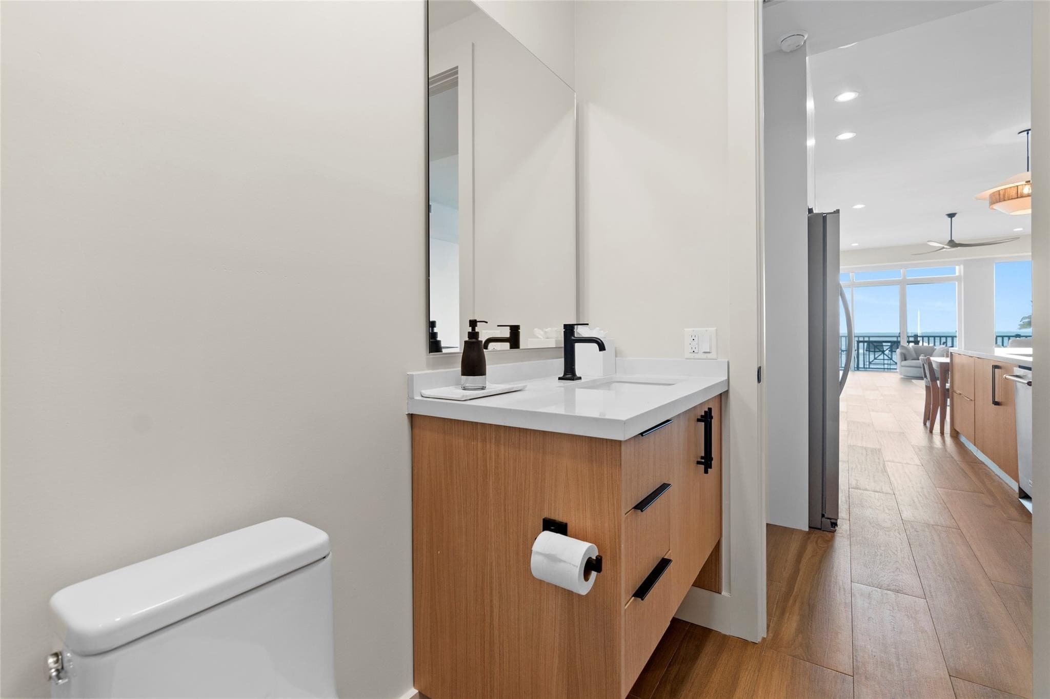 Powder room with a floating wood vanity, black faucet and hardware, and an open pocket door revealing the wood-floored living area and bay view beyond.
