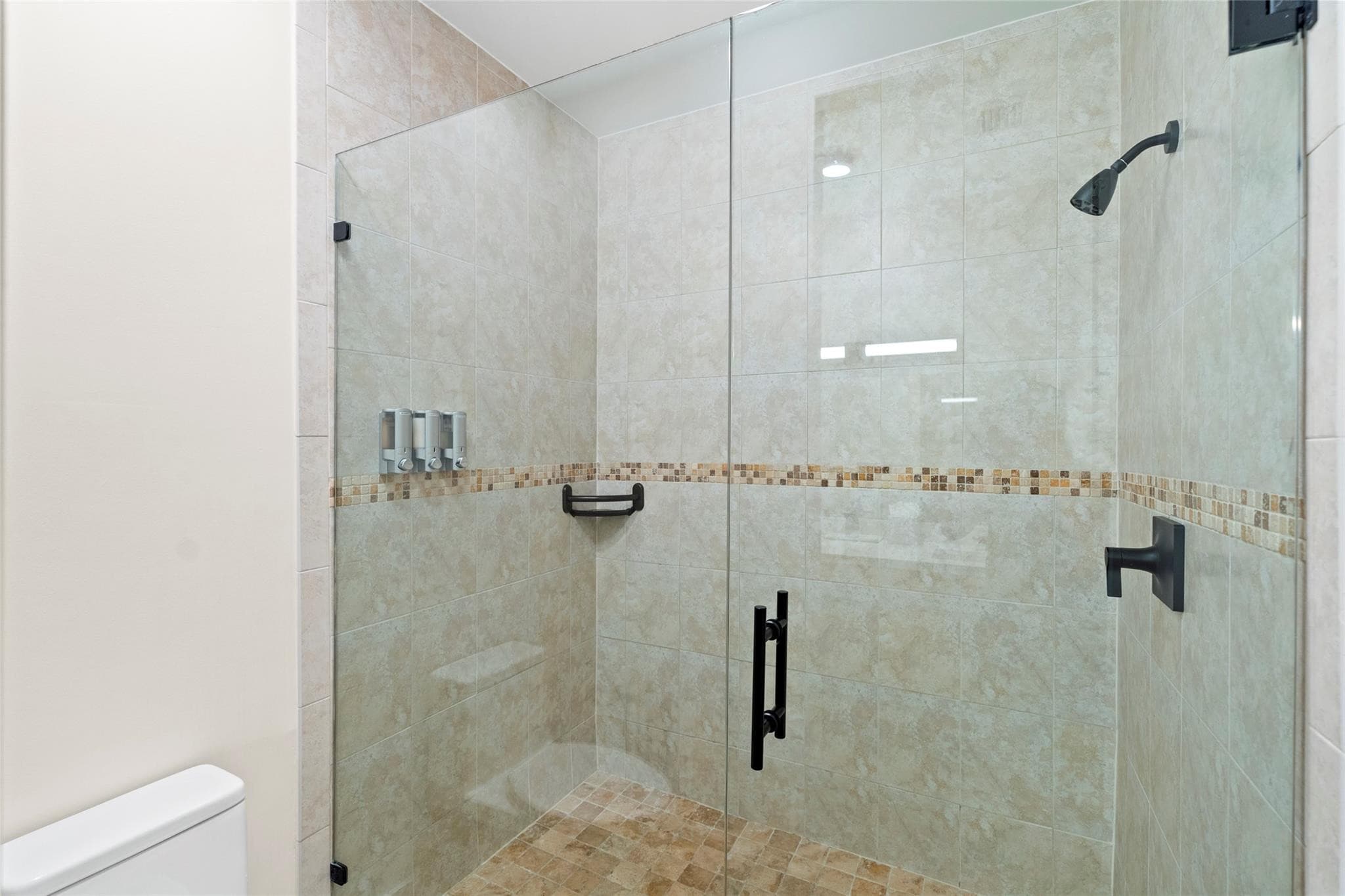 Frameless glass walk-in shower with warm beige tile, mosaic accent stripe, black shower fixtures, and built-in dispensers.