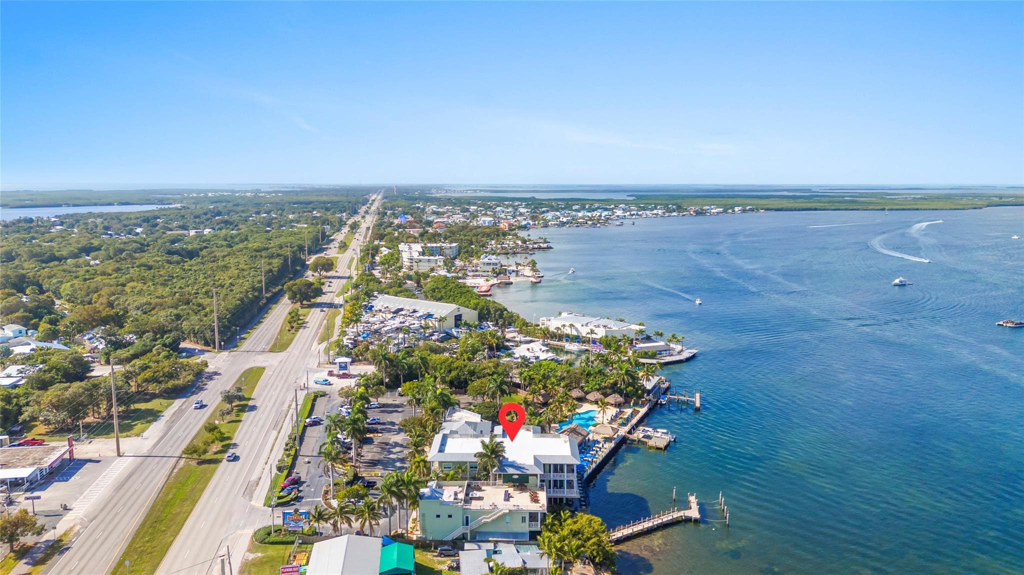 Sweeping aerial along the island highway and bayside coastline, with a red pin marking the waterfront property, its rooftop deck, pool, and dock jutting into teal water.