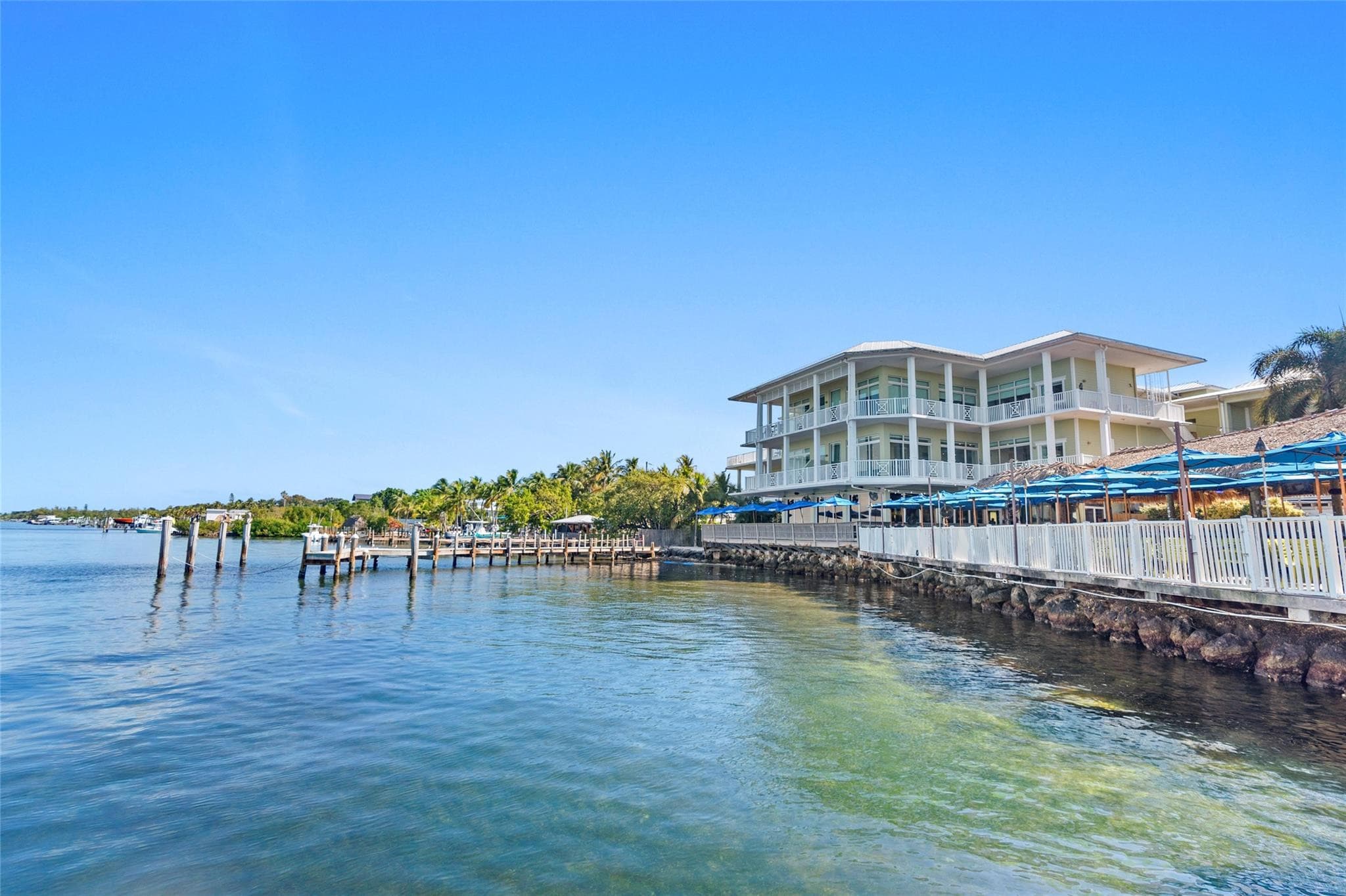 View from the water of the pale-yellow three-story residence building with white balconies and metal roof, fronted by a rock seawall, docks, and rows of blue umbrellas.