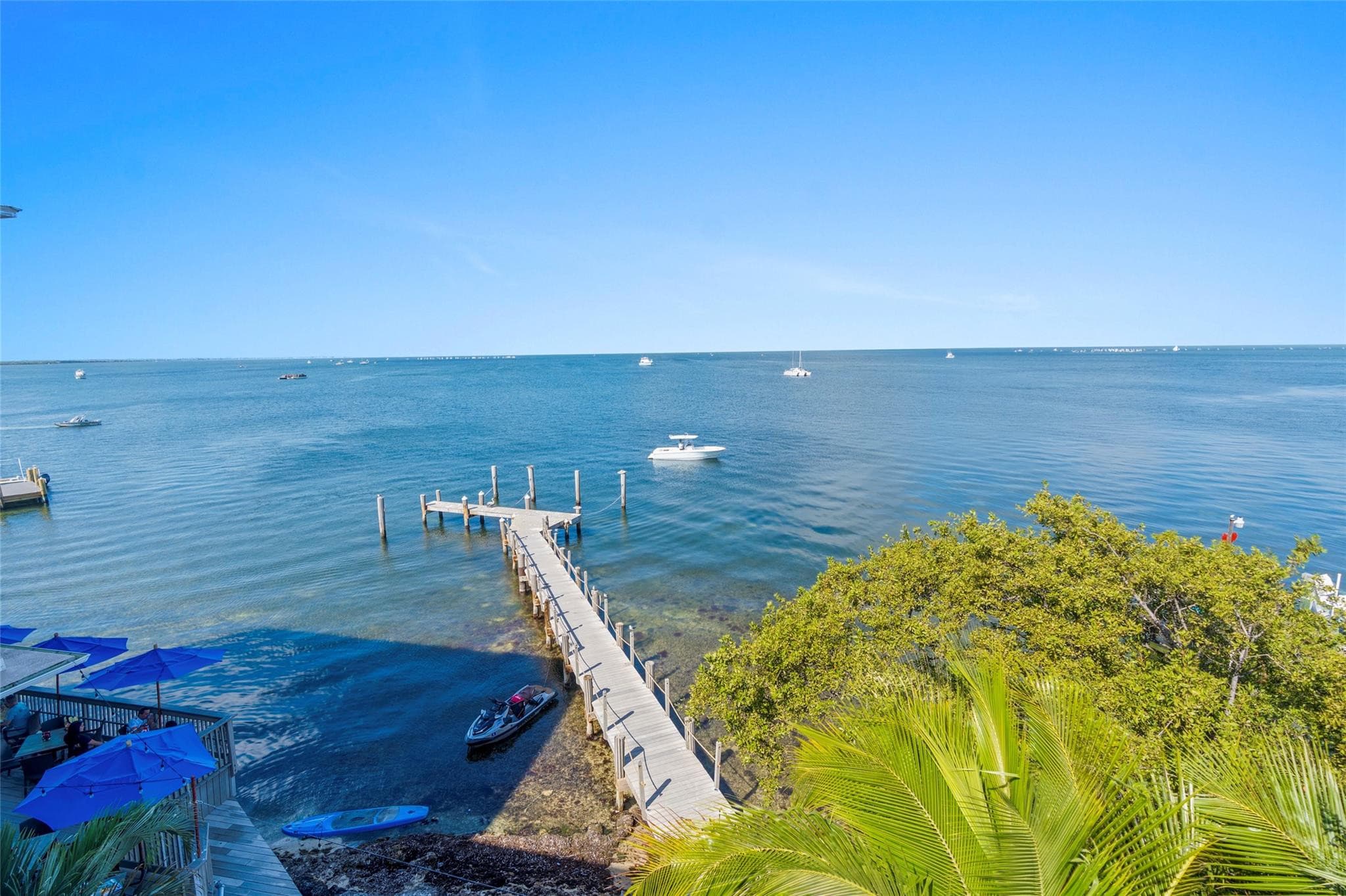 Elevated view of a long wooden boardwalk dock extending into clear bay water, with a jet ski and paddleboard at the shoreline and boats on the horizon.