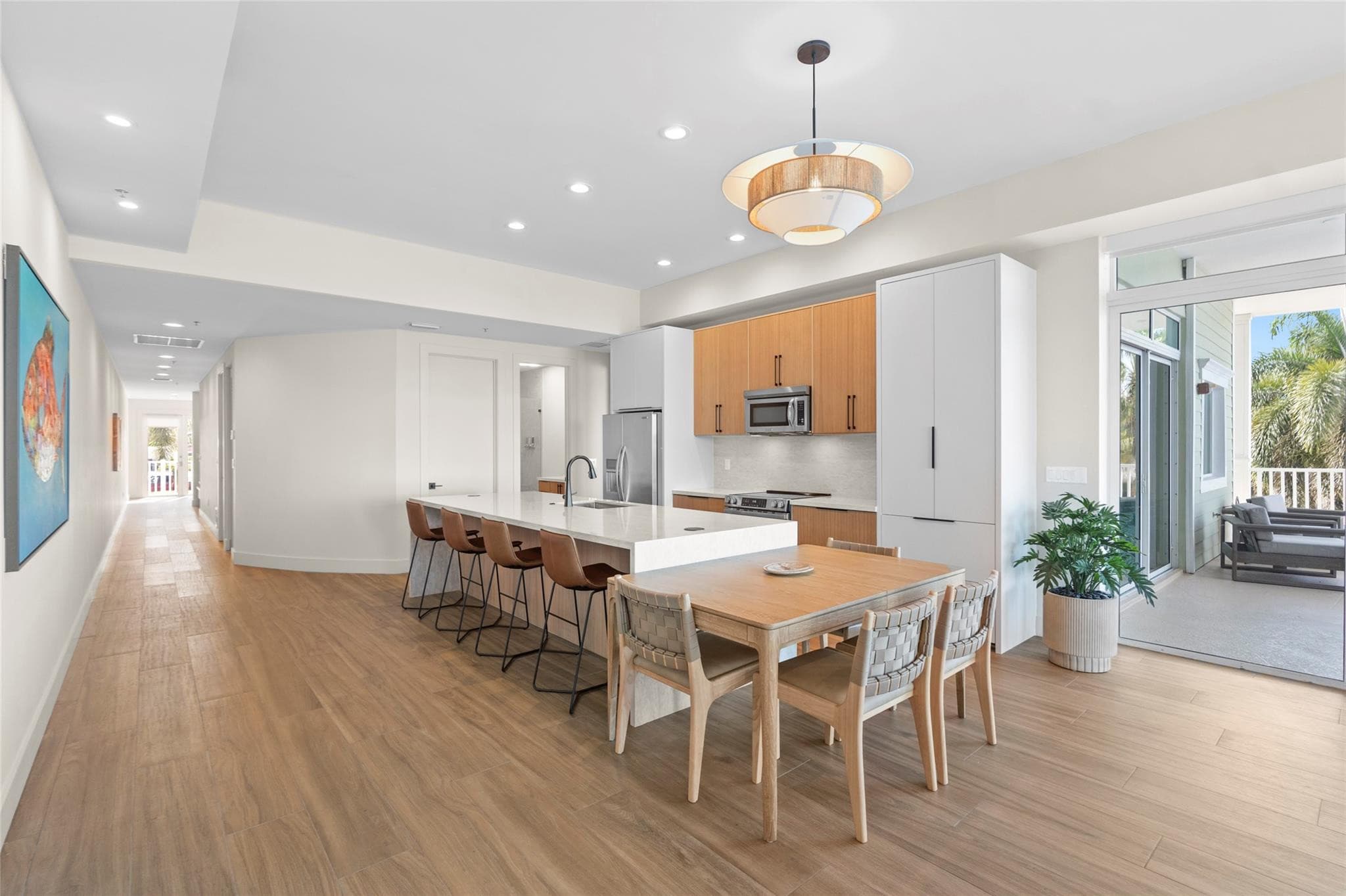 Open kitchen and dining area with a white quartz island, leather barstools, light-wood dining table with woven chairs, rattan pendant light, and balcony access.