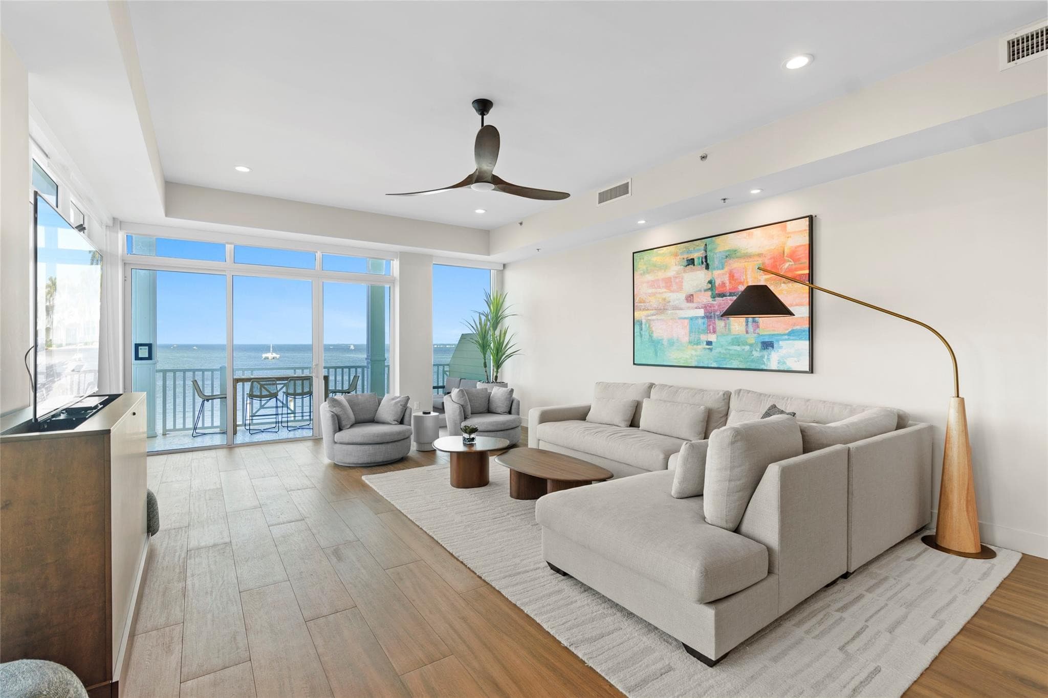 Living room with a large beige sectional and chaise, two swivel chairs, brass arc lamp, colorful abstract canvas, and glass doors framing the bay horizon.