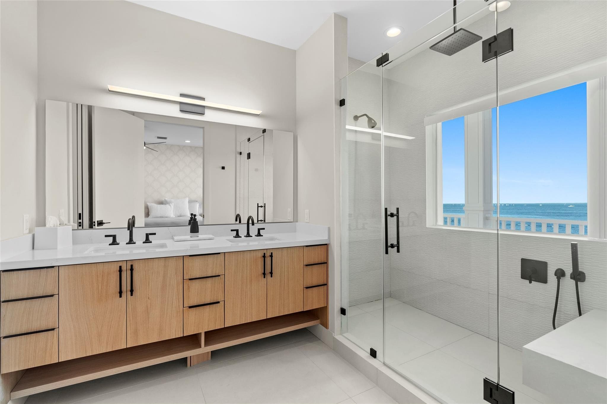 Primary bathroom with a white-oak double vanity, matte-black fixtures, and a glass walk-in shower lit by a picture window overlooking the water.