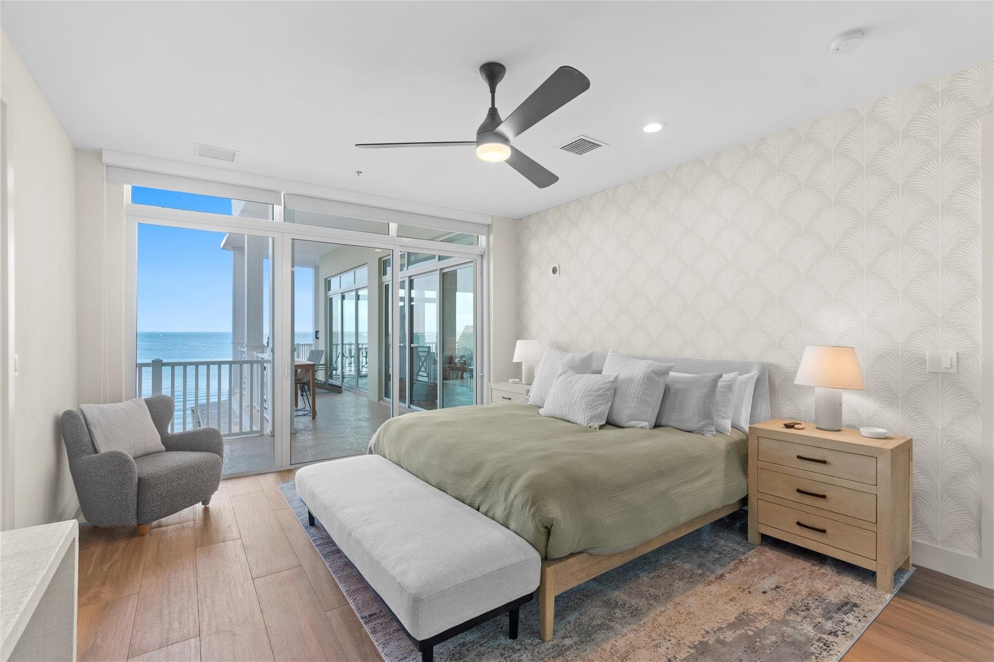 Primary bedroom with a king bed in sage-green linens, palm-pattern accent wallpaper, a gray armchair, and sliding doors opening to a balcony over the bay.