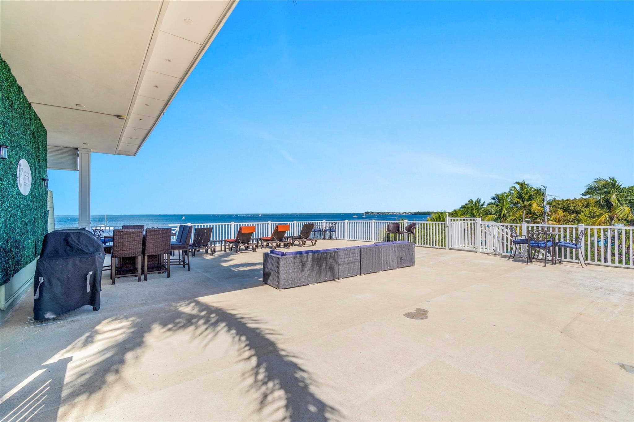 Vast rooftop sun deck with a covered grill, wicker dining sets, orange-cushioned loungers, and a long sectional along white railings overlooking the bay.
