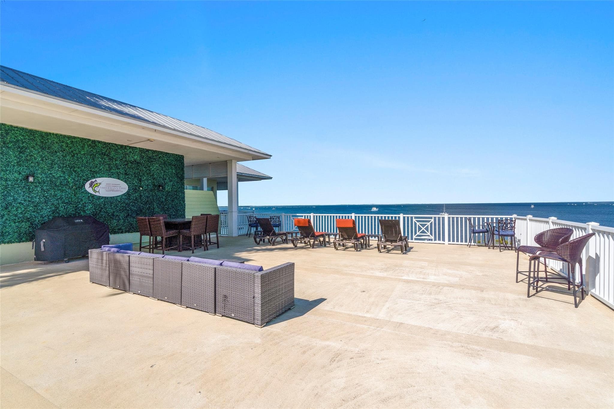 Rooftop terrace with a green hedge wall bearing the club's sailfish emblem sign, covered grill, wicker sectional with navy cushions, and chaise lounges facing the water.