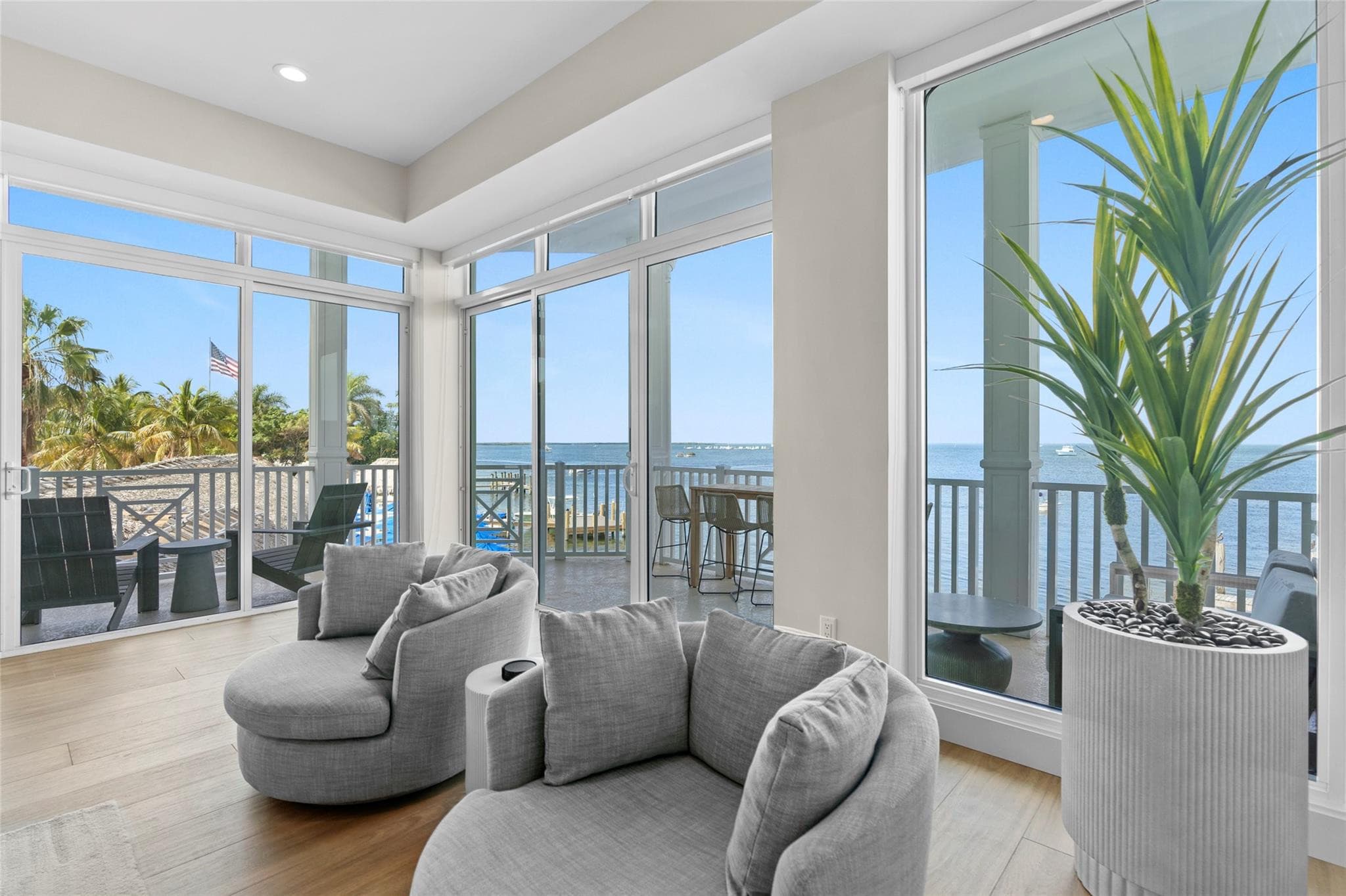 Corner sitting nook with two plush gray swivel chairs and a tall potted plant, surrounded by wraparound glass doors opening to balconies over the bay.