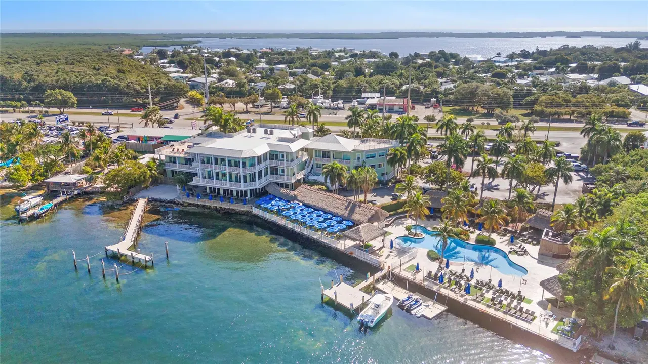 Aerial view of the bayfront residence club showing the white three-story building, free-form pool, thatched tiki huts, blue umbrellas, docks, and palm-lined grounds along the turquoise bay.