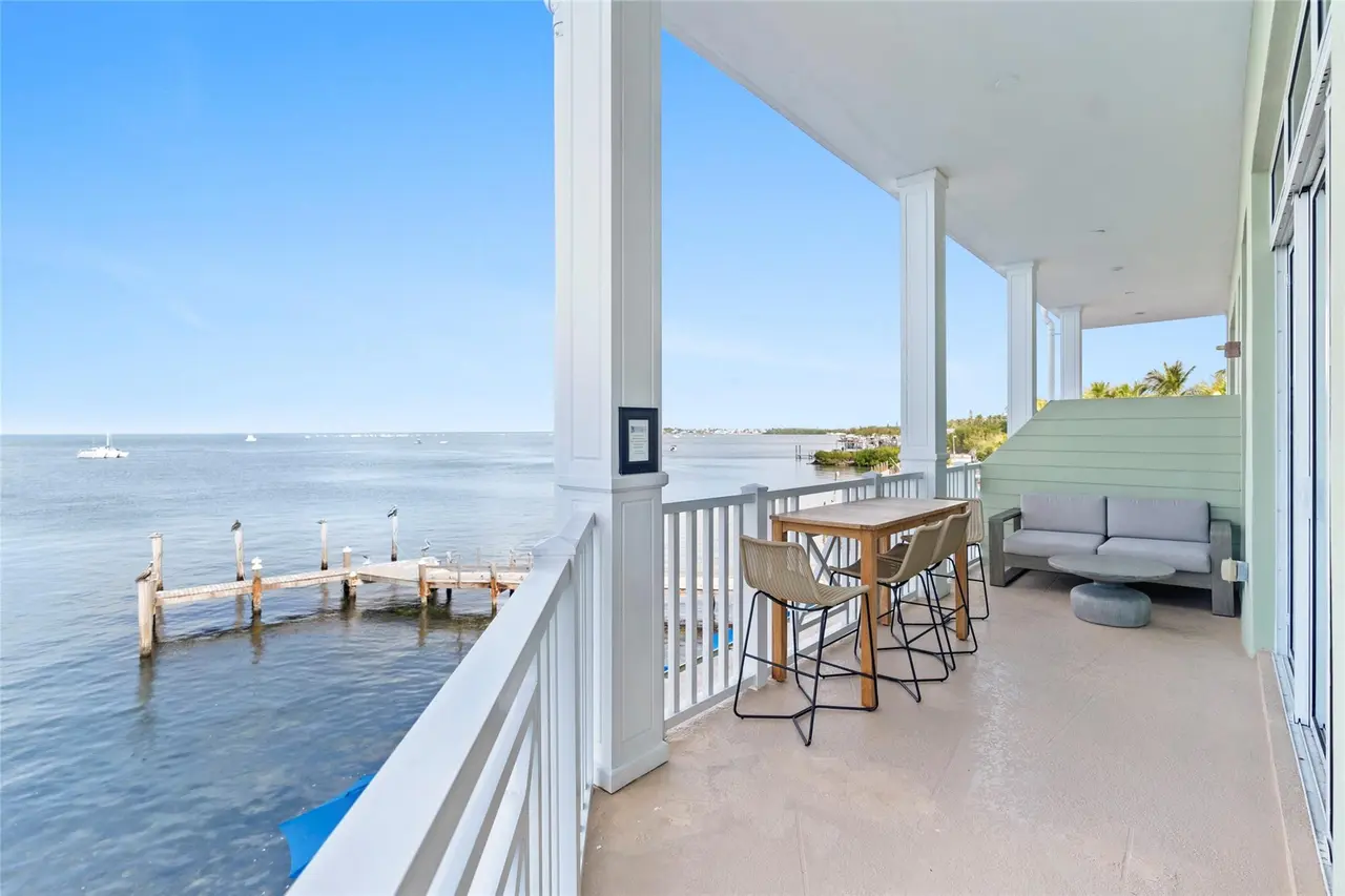 Covered balcony with white columns and railings, a teak bar-height table, wicker stools, and a gray outdoor sofa overlooking weathered dock pilings and calm bay water.