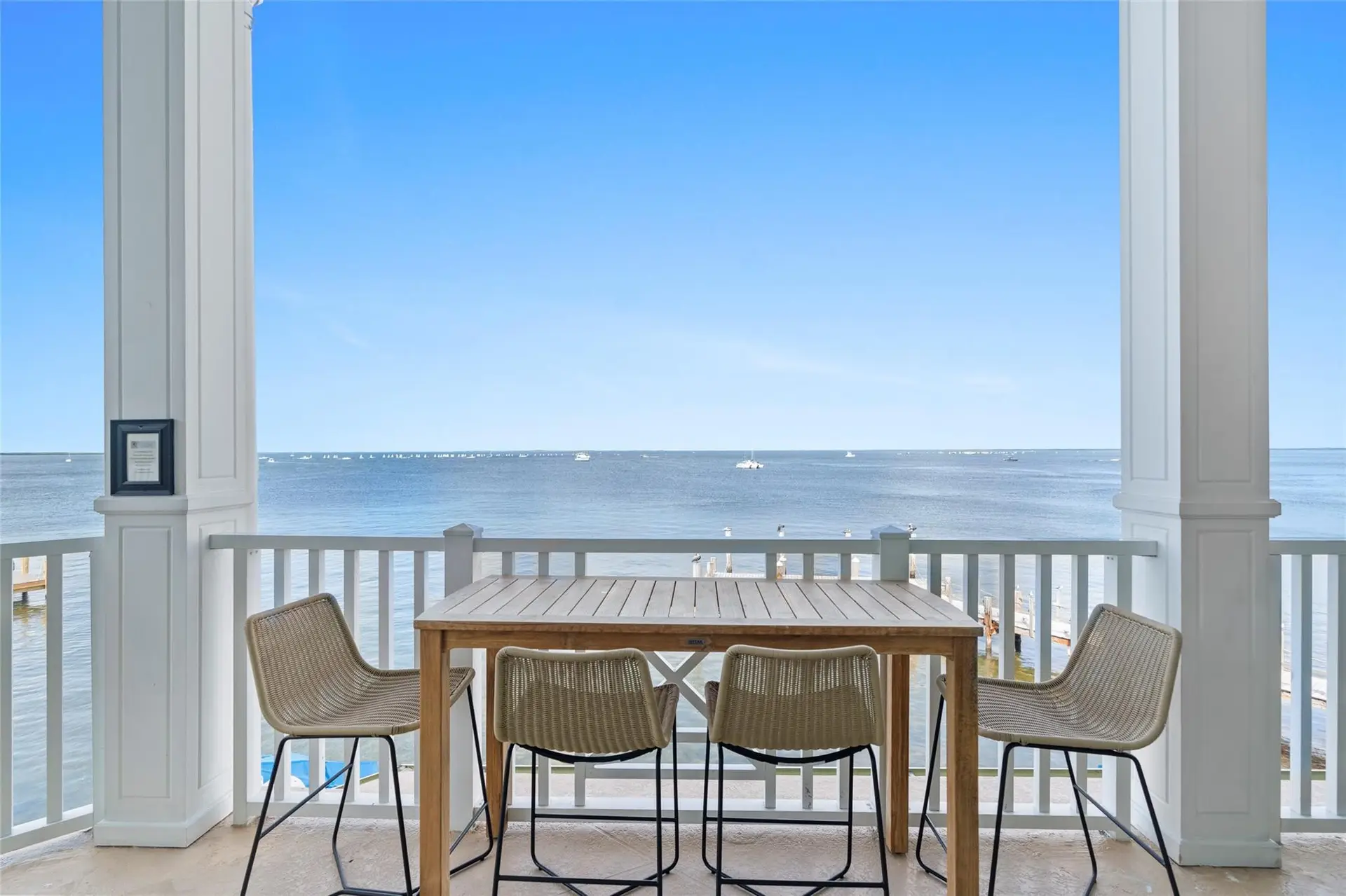 Teak bar-height table with four woven counter chairs on a columned balcony facing an unbroken horizon of calm bay water dotted with distant sailboats.
