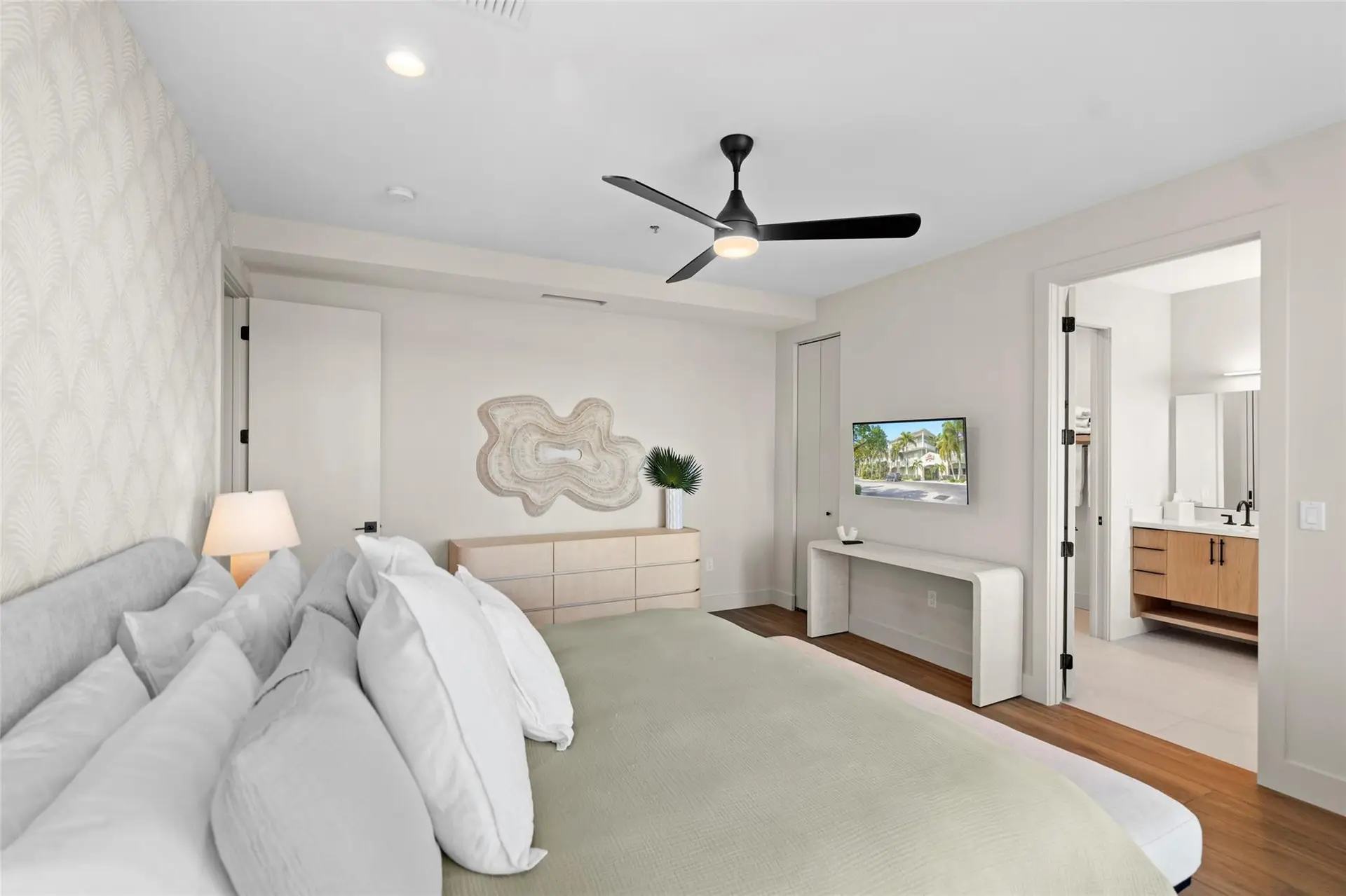 Neutral-toned bedroom with layered white pillows, a light-wood dresser under sculptural wall art, wall-mounted TV, and a doorway to the en-suite vanity.
