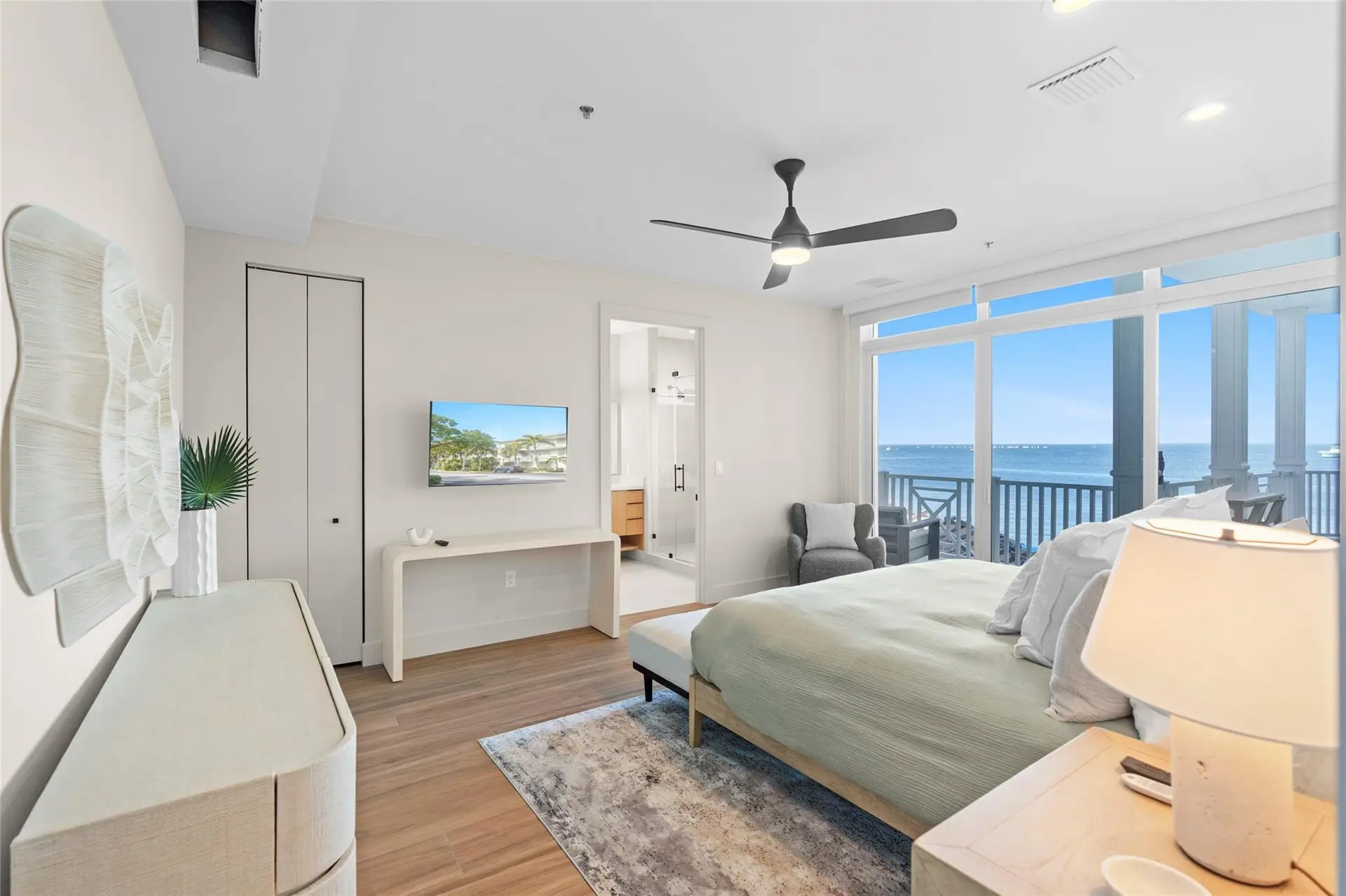 Spacious bedroom with woven wall art, a white console table with TV, en-suite bath, and floor-to-ceiling glass opening to a balcony over the open water.