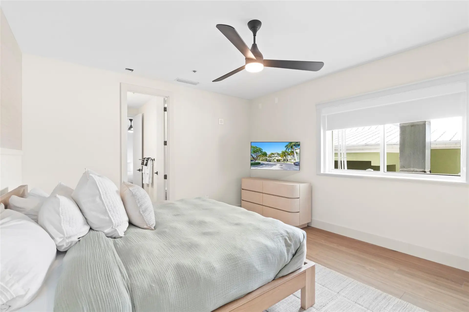 Guest bedroom with sage bedding and stacked white pillows, a light-wood dresser below a wall-mounted TV, ceiling fan, and a wide window with roller shade.