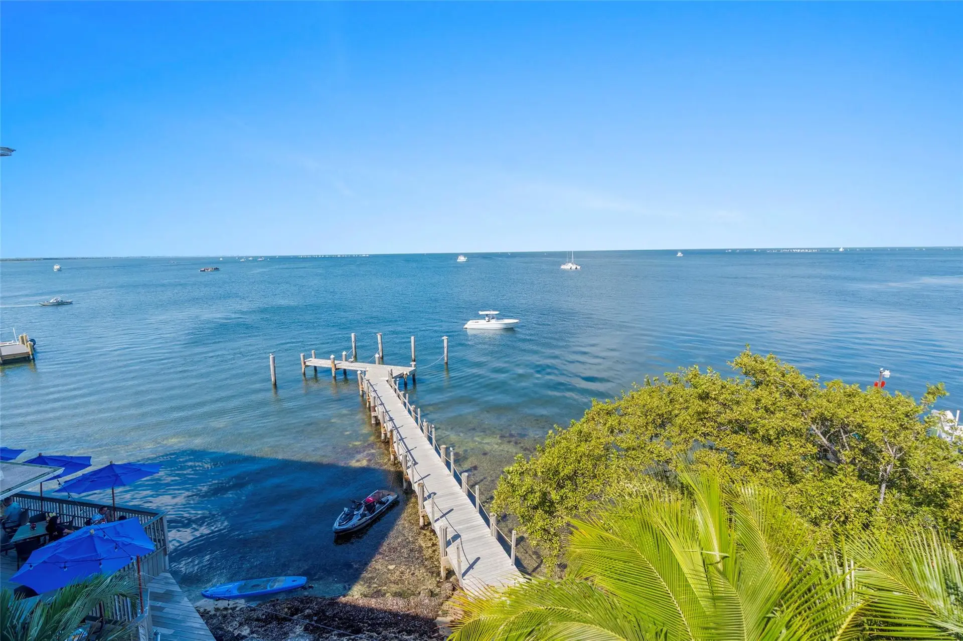 Elevated view of a long wooden boardwalk dock extending into clear bay water, with a jet ski and paddleboard at the shoreline and boats on the horizon.