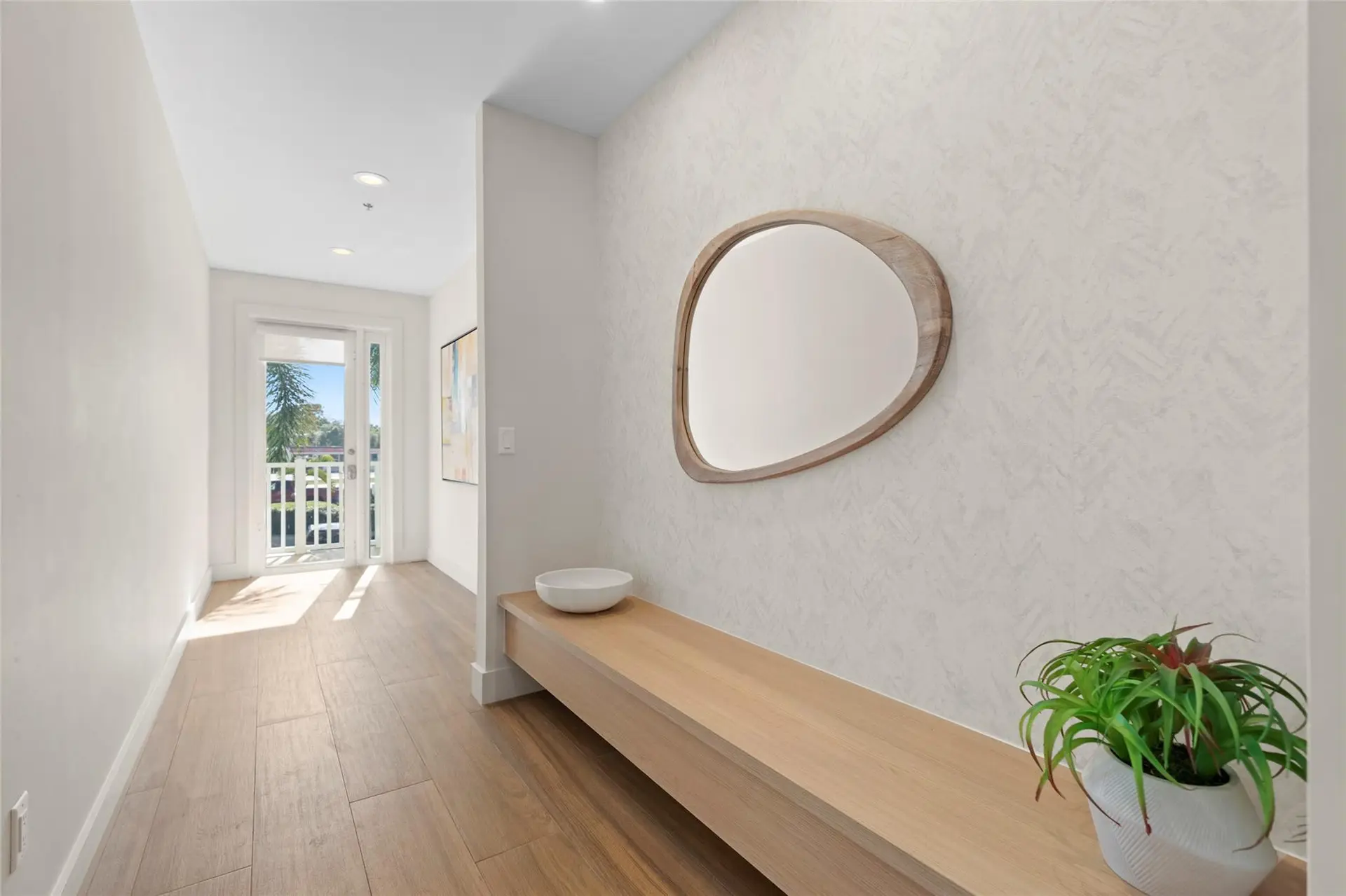 Bright foyer with a long floating wood bench, sculptural mirror on textured wallpaper, potted plant, and a glass French door letting in palm-filtered sunlight.