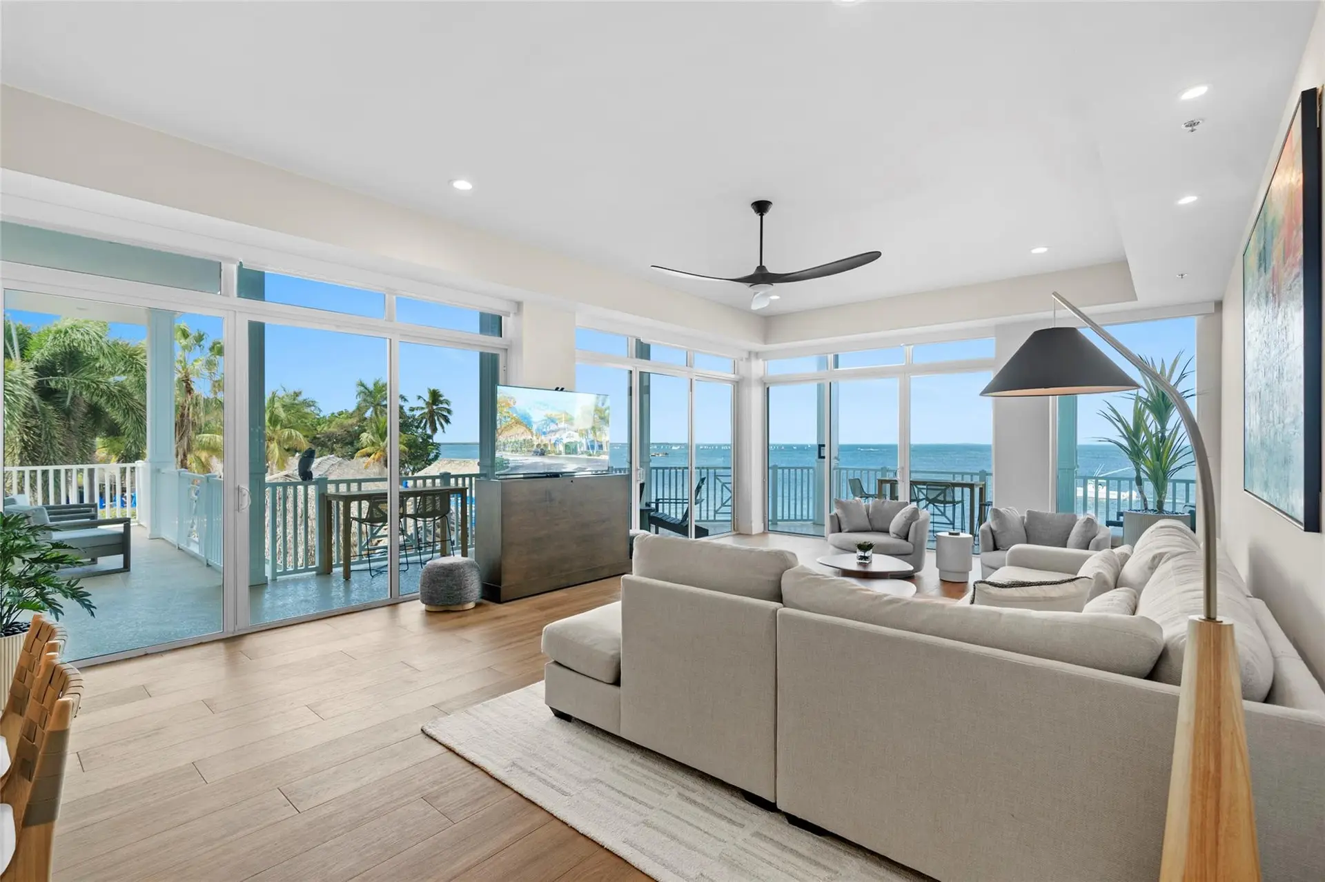 Corner living room wrapped in floor-to-ceiling windows and balcony doors with panoramic bay views, a beige sectional, arc floor lamp, and pop-up TV console.