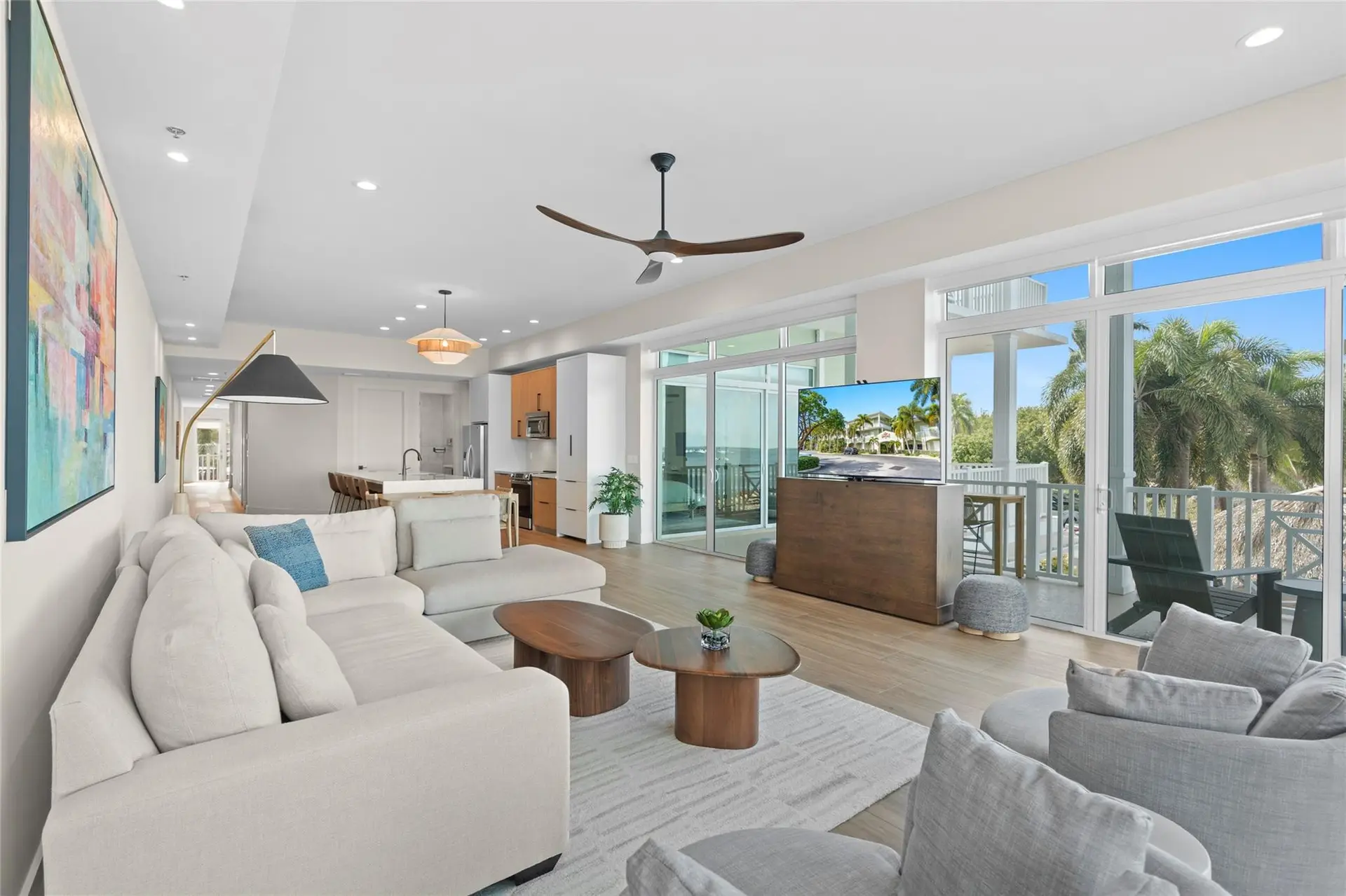 Bright living room with a cream sectional, walnut coffee tables, colorful abstract art, a TV lift cabinet, and sliding glass doors to a covered balcony framed by palms.
