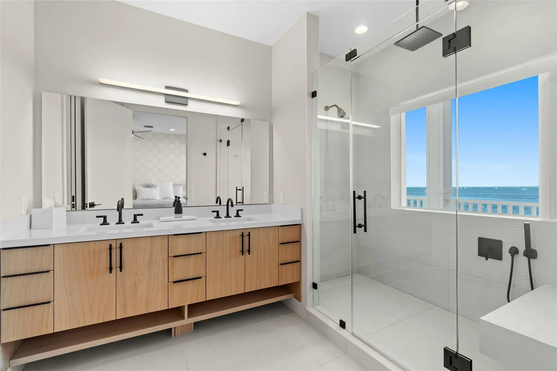 Primary bathroom with a white-oak double vanity, matte-black fixtures, and a glass walk-in shower lit by a picture window overlooking the water.