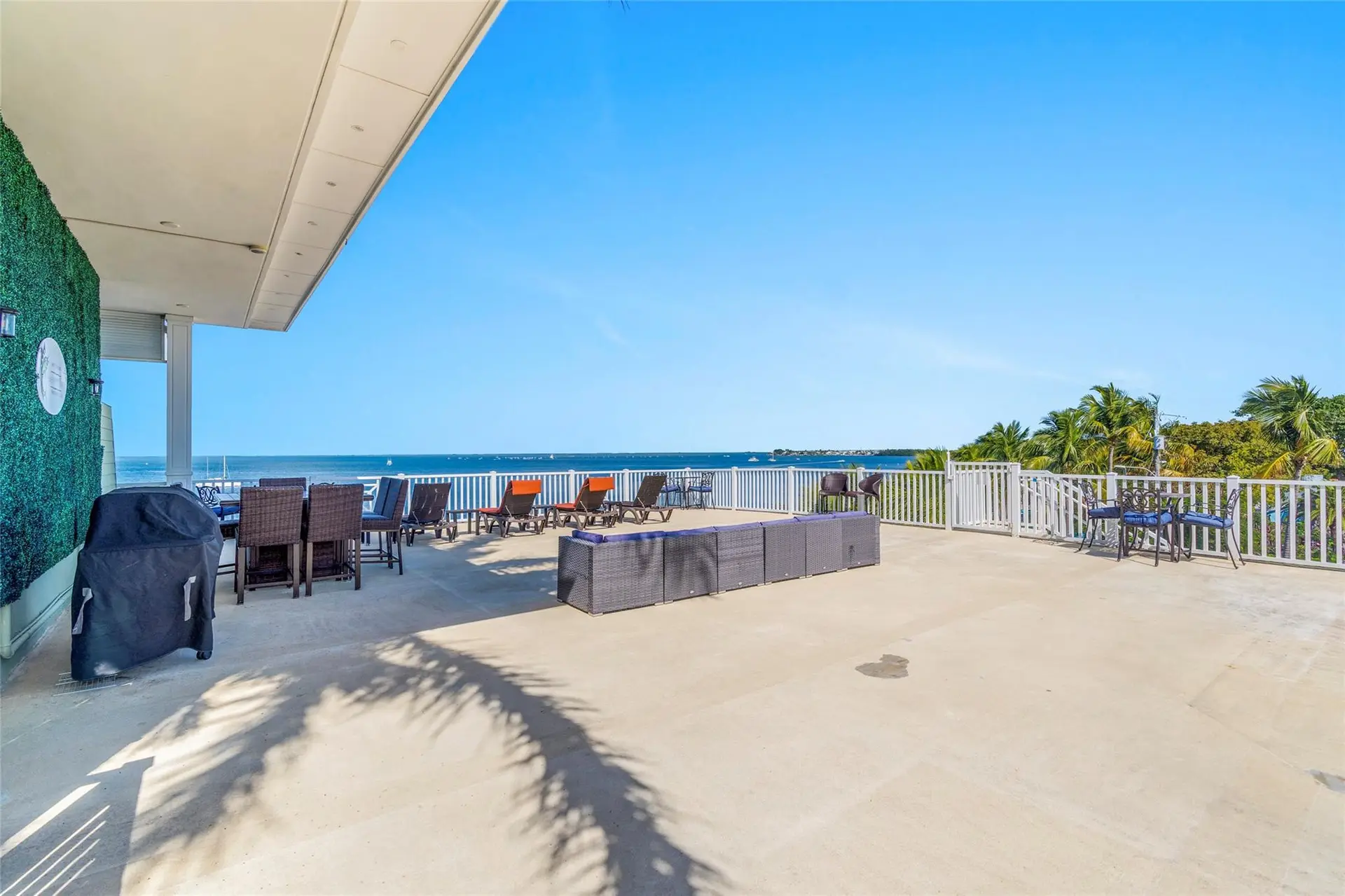 Vast rooftop sun deck with a covered grill, wicker dining sets, orange-cushioned loungers, and a long sectional along white railings overlooking the bay.