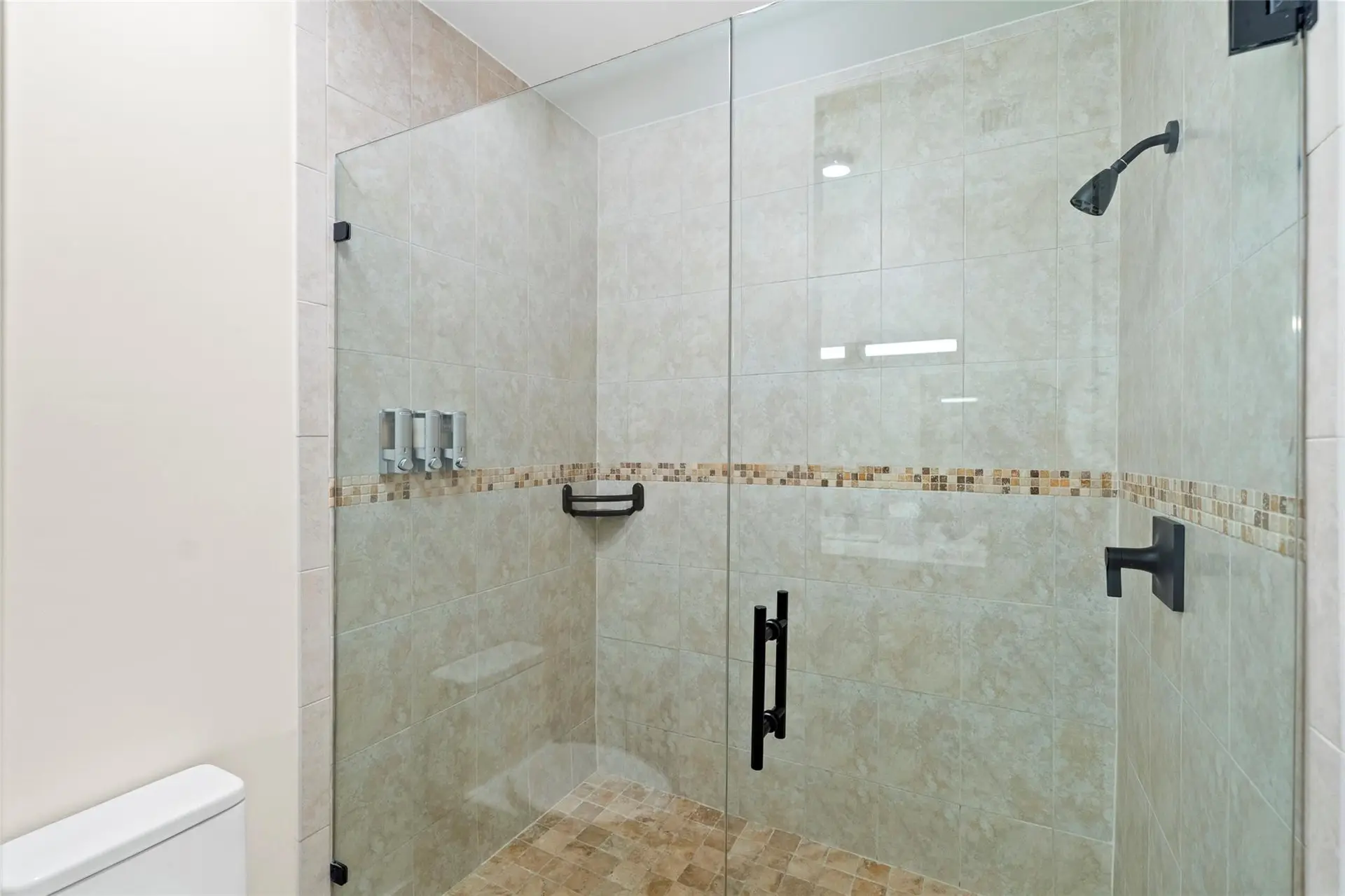 Frameless glass walk-in shower with warm beige tile, mosaic accent stripe, black shower fixtures, and built-in dispensers.